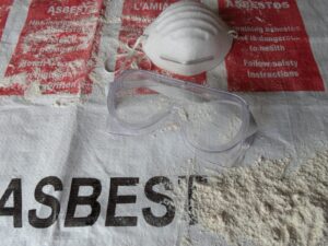 Goggles and a protective mask on top of a sign with asbestos hazard warnings in multiple languages.