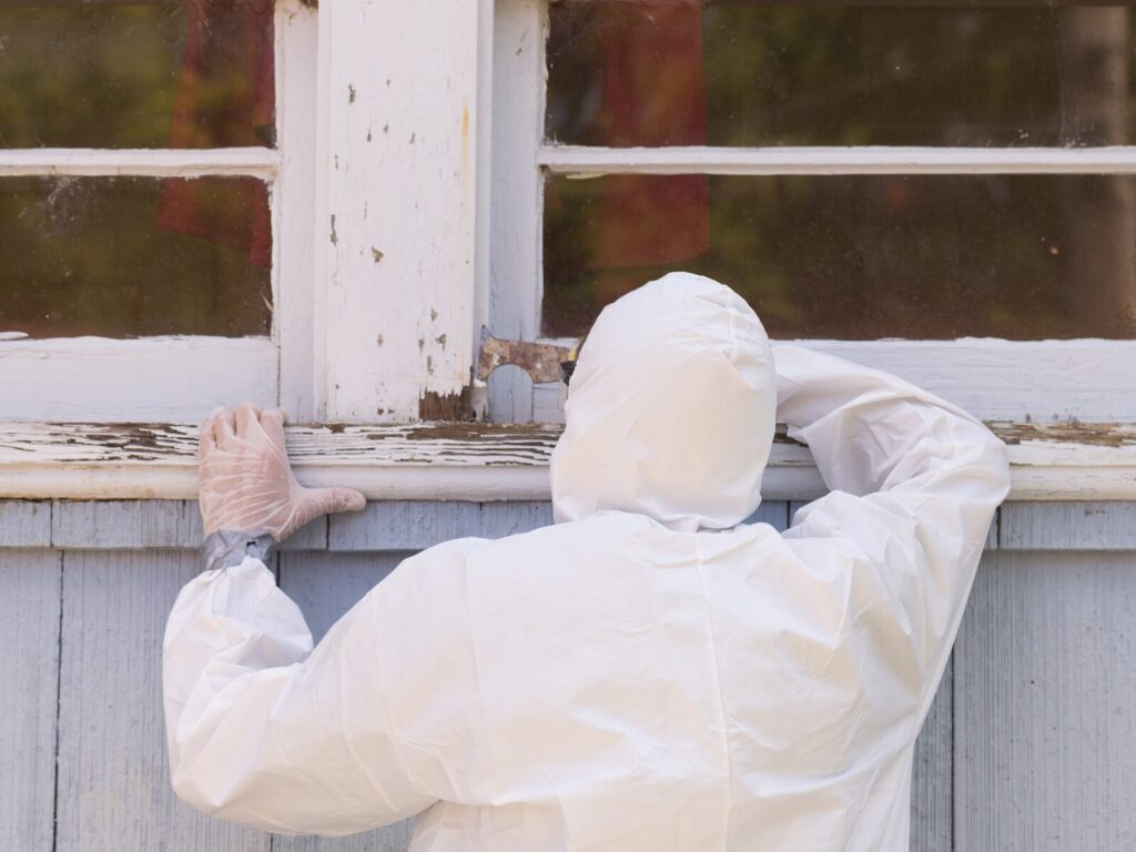 A man wearing protective clothing and gloves scrapes a paint sample from a window pane.
