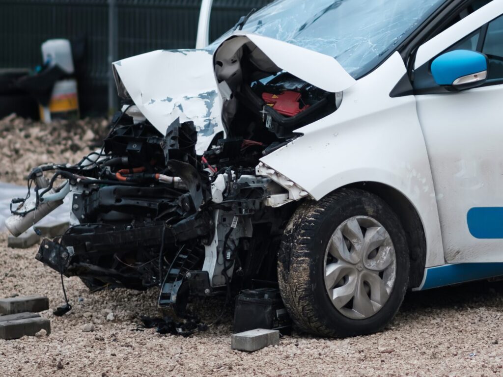 The front end of an electric vehicle after a collision. The grille and fender are crushed, but the vehicle's battery has resisted the impact.