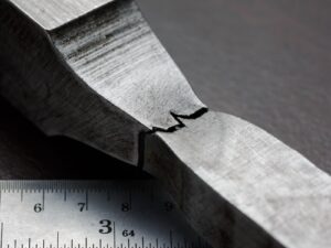 Metal specimen after tensile testing. The specimen has a crack and signs of stretching. It rests on top of a ruler.