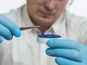 Chemist holds a tiny plastic pellet between tweezers in one hand. In his other hand, he holds a shallow bowl of other plastic pellets.