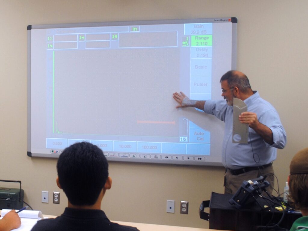 Expert holds a metal component up to a classroom for inspection and gestures to a screen